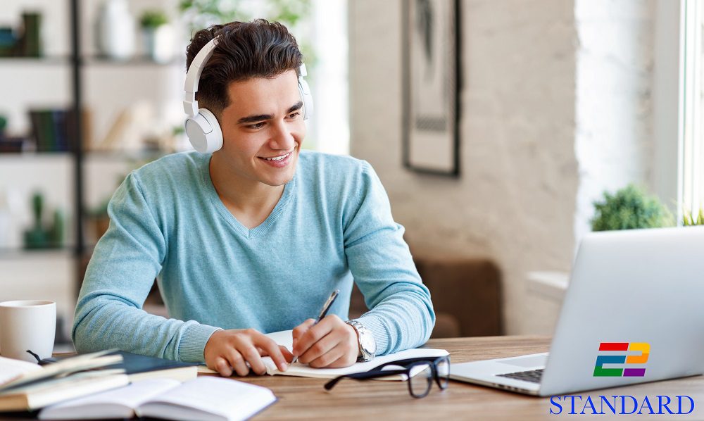 Happy ethnic guy in headphones smiling and writing in notebook while sitting at table and listening to teacher during online lecture at home